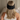 Back view of woman with long hair wearing a black silk sleep mask with comfort wrap, seated on a bed.