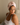 Woman wearing a light pink ZILK silk hair bonnet, seated on a bed and looking to the side in soft natural light