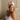 Woman wearing a light pink ZILK silk hair bonnet, seated on a bed and looking to the side in soft natural light